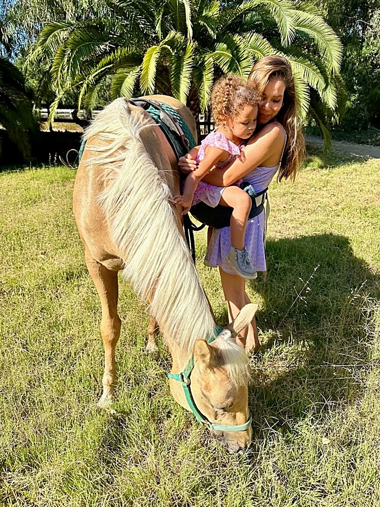 A therapist holds a young child close beside a gentle palomino horse grazing in the grass