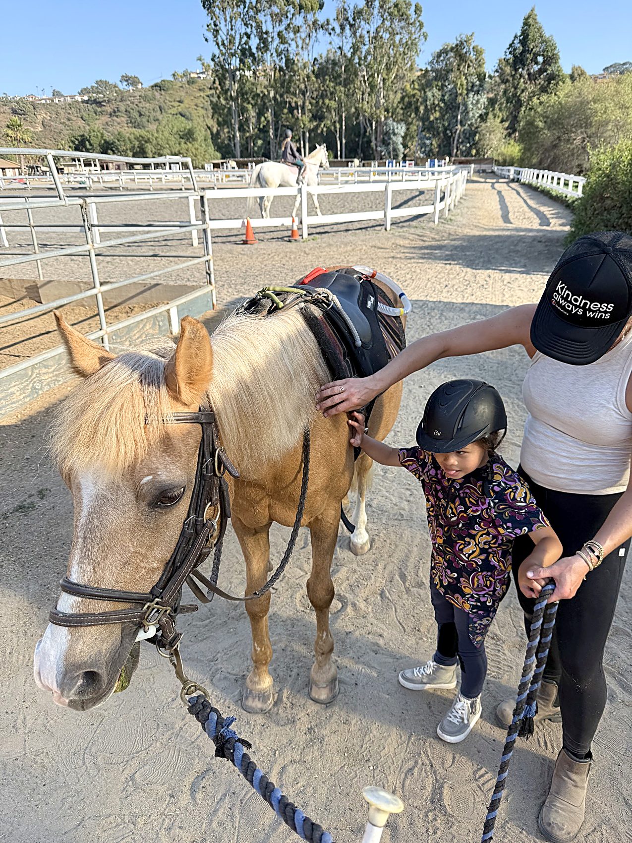 A young child reaches up to touch a pony's muzzle while an instructor stands alongside in a sunlit arena
