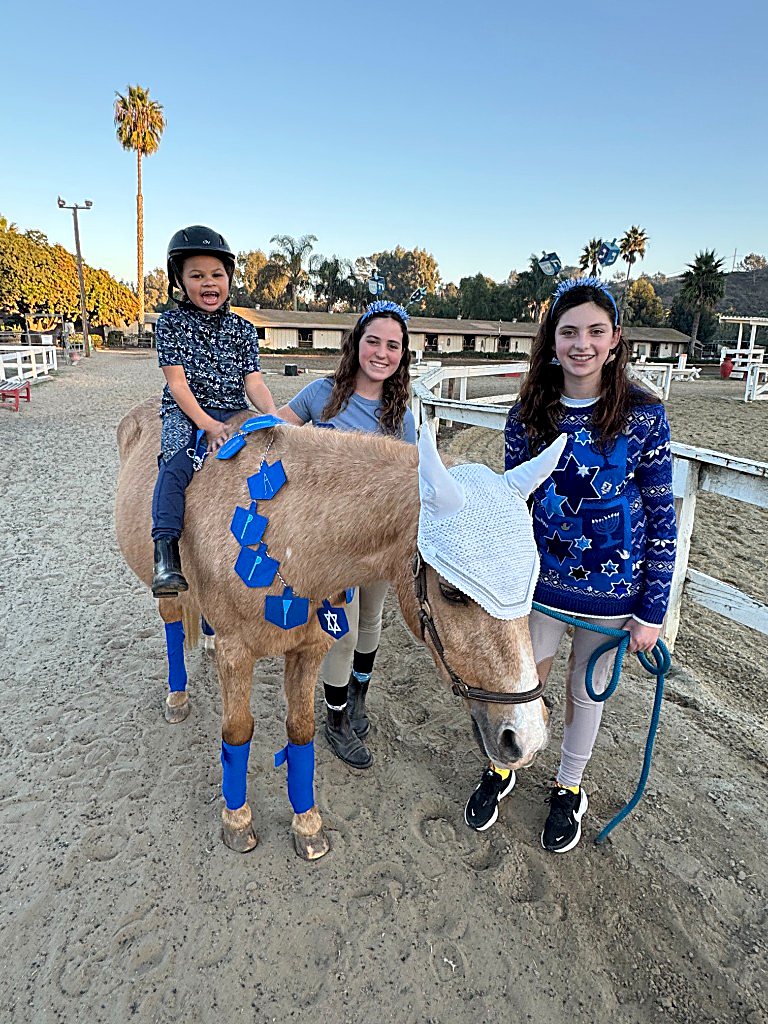 Two teenage volunteers smile beside the palomino pony as a young rider sits in the saddle
